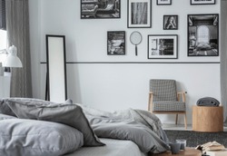 Gallery of black and white photos on wall of stylish bedroom interior with mirror, retro armchair and wooden table with blanket