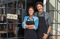 Two cheerful small business owners smiling and looking at camera while standing at entrance door. Happy mature man and mid woman at entrance of newly opened restaurant with open sign board.