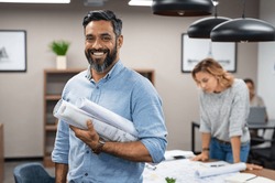 Portrait of multiethnic architect with blueprints in creative office. Mature middle eastern contractor holding roll of architectural projects while looking at camera. Happy latin man in casual.