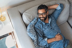 Mature man resting on sofa and thinking about the future. Hispanic man lying on couch and looking away. Smiling casual guy relaxing and daydreaming on sofa at home.