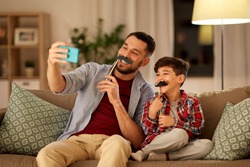 people, family and technology concept - happy father and little son with mustaches party props taking selfie by smartphone sitting on sofa at home in evening