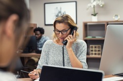 Mature businesswoman talking on phone in office. Casual business woman talking on landline phone in a creative agency. Beautiful blonde receptionist with eyeglasses taking client details over phone.