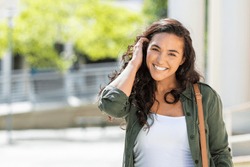 Happy young beautiful woman walking on the street. Portrait of cheerful university student looking at camera while adjusting curly hair with copy space. Latin stylish girl smiling while standing.