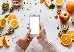 Female hands holding smartphone on healthy food background, woman using phone search mobile apps with diet nutrition plan cooking, vegan fruit granola seeds on white table, top view, mock up screen