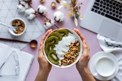 Female hand holding healthy breakfast bowl over work table background concept enjoy detox morning meal with laptop milk, woman eat natural granola nutrition detox food in home office, top view
