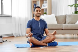 fitness, meditation and healthy lifestyle concept - indian man in earphones listening to music on smartphone and meditating in lotus pose at home