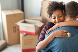 Young african woman holding home keys while hugging boyfriend in their new apartment after buying real estate. Lovely girl holding keys from new home and embracing man. Couple around cardboard boxes.