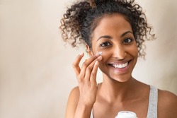 Smiling african girl with applying facial moisturizer while holding jar and looking at camera. Portrait of young black woman applying cream on her face isolated on beige background with copy space.