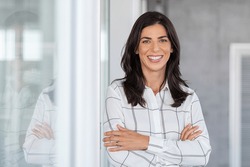 Portrait of middle aged businesswoman in modern office looking at camera. Confident business woman with arms crossed standing while leaning against glass wall. Proud woman smiling in with copy space.