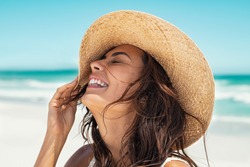 Close up face of young stylish woman wearing straw hat at beach. Happy tanned latin woman laughing during summer holiday. Beautiful fashionable girl relaxing at beach while holding large brim for wind