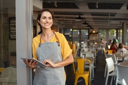Portrait of a happy waitress standing at restaurant entrance holding digital tablet. Happy mature woman owner in grey apron standing at coffee shop entrance leaning while looking away with copy space.