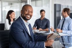 Proud mature black businessman smiling with colleagues sitting in a board room. Portrait of happy successful executive with team working in background while looking at camera during meeting. Success.