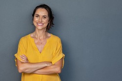 Confident mature woman with crossed arms in casual clothing with copy space. Successful smiling woman with big grin looking at camera. Beautiful positive businesswoman standing against grey background