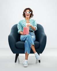 comfort, people and furniture concept - portrait of happy smiling young woman in 3d movie glasses eating popcorn from striped bucket sitting in modern armchair over grey background