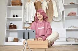 Happy hipster gen z teen girl fashion social media channel blogger with pink hair wear hoodie recording unboxing vlog on phone video camera tutorial sit on floor with online store clothes order box.