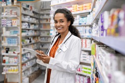 Happy friendly multiethnic pharmacist doing inventory in a provided and modern pharmacy while looking at camera. Portrait of smiling african doctor woman working in drugstore with digital tablet.