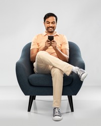 people, technology and furniture concept - happy smiling young indian man with smartphone sitting in chair over grey background