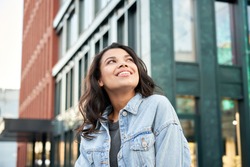 Confident happy beautiful young hipster African American woman wearing denim jacket looking up standing on city street outdoors dreaming, thinking or good future on urban buildings background.