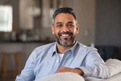 Portrait of happy mid adult man sitting on sofa at home. Handsome latin man in casual relaxing on couch and smiling. Cheerful indian guy looking at camera. 