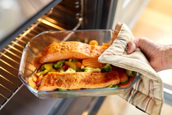 food cooking, culinary and people concept - young woman with potholder taking baking dish with salmon fish and vegetables out of oven at home kitchen