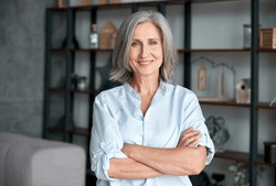 Smiling confident stylish mature middle aged woman standing at home office. Old senior businesswoman, 60s gray-haired lady executive business leader manager looking at camera arms crossed, portrait.