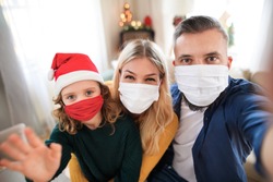 Family with small daughter indoors at home at Christmas, taking selfie with face masks.