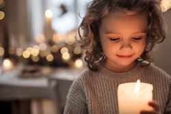 Cheerful small girl indoors at home at Christmas, holding candle.
