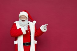 Funny happy excited old bearded Santa Claus face wearing costume looking at camera showing pointing fingers aside advertising Christmas promotion, New Year xmas discount ad isolated on red background.
