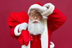 Surprised shocked funny old Santa Claus face wearing costume holding sack bag with Christmas gifts presents looking at camera advertising promotion, New Year xmas discount isolated on red background.