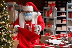 Old funny bearded Santa Claus wearing costume, face mask, packing presents gift boxes in sack bag preparing for xmas eve in workshop. Merry Christmas Covid 19 coronavirus safe delivery concept.