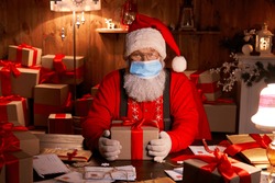Old funny bearded Santa Claus wearing face mask, holding gift box preparing for xmas eve sitting at cozy home table late in night with presents. Merry Christmas Covid 19 coronavirus safe delivery.