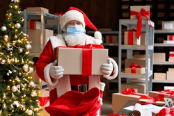 Old bearded Santa Claus wearing costume, face mask, holding present gift box packing in sack bag preparing for xmas eve in workshop. Merry Christmas Covid 19 coronavirus safe delivery concept.