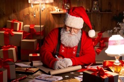 Happy old kind bearded Santa Claus wearing hat, glasses, writing on wish list, working on Christmas eve sitting at cozy home workshop table late with presents, tree and candles preparing for holidays.