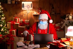 Old bearded Santa Claus wearing face mask, holding gift box on xmas eve sitting at cozy home table late in night using laptop computer. Merry Christmas Covid 19 coronavirus social distance concept.