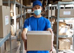 Male courier worker wearing uniform and face mask holding parcel box giving package to camera standing in stock warehouse, portrait. Fast express safe delivery during covid 19 pandemic concept.