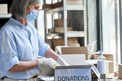 Mature female warehouse worker volunteer wearing face mask working in shipping delivery charitable stock organization packing donations box. Covid 19 coronavirus donating and volunteering concept.