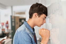 Disappointed university student leaning on whiteboard with closed eyes. Tired high school guy feeling failure in classroom. Side view of sad young college student trying to solve math problem.