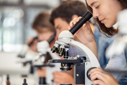 Close up of young woman seeing through microscope in science laboratory with other students. Focused college student using microscope in the chemistry lab during biology lesson.