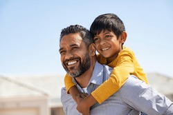 Happy middle eastern child enjoying ride on father back outdoor. Smiling dad giving piggyback ride to son on street while looking away with copy space. Indian cheerful man carrying on shoulder kid.