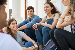 Group of happy young friends sitting in college campus and talking. Cheerful group of  smiling girls and guys feeling relaxed after university exam. Excited millenials laughing and having fun outdoor.