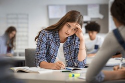 Anxious young woman with hand on head feeling tired while studying at school. College student suffering from headache in classroom. Troubled and stressed girl doing exam that doesn’t know the answers.