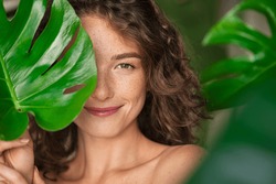 Close up face of beautiful young woman covering her face by green monstera leaf while looking at camera. Portrait of beauty woman with natural makeup and freckles standing behind big green leaves.
