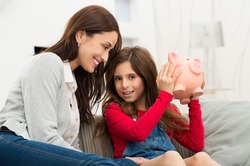 Smiling Mother Looking At Her Daughter Sitting On Couch Holding Piggybank