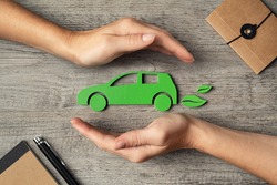 Close up of hands of woman protecting icon of green eco-friendly car over wooden table. Top view of woman hands protecting electric car with care, mobility. Car insurance and green automotive concept.