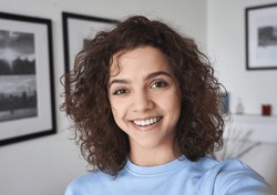 Smiling cute cheerful young pretty hispanic latin woman looking at camera standing alone at home. Happy positive beautiful 20s girl female model posing indoors, close up face headshot portrait.
