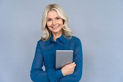 Happy elegant middle aged older professional business woman leader, consultant manager, looking at camera holding digital tablet isolated on grey wall advertising corporate online e learning webinars.