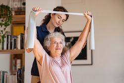 Young trainer helping senior woman with resistance band. Lovely physiotherapy health worker exercise with old woman at home. Happy smiling senior woman using rubber band to improve her muscle tone.