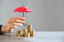 Young woman hand holding small red umbrella over pile of coins on table. Close up of stack of coins with female hands holding umbrella for protection. Financial safety and investment concept.  