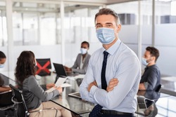 Portrait of mid businessman wearing protective face mask while his team working in background. Successful entrepreneur in conference room leaning over table and looking at camera with copy space.