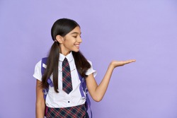 Smiling indian schoolgirl, preteen kid smiling looking at copy space holding hand. Advertising latin junior student standing isolated on violet lilac background. Education, school concept.